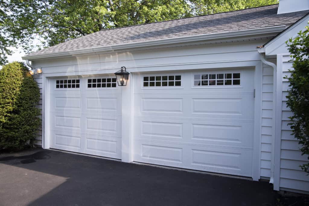 Two white garage doors with rectangular windows along the top, set in a white house exterior. A black lantern-style light fixture is mounted between the doors. Green trees and shrubs surround the area.