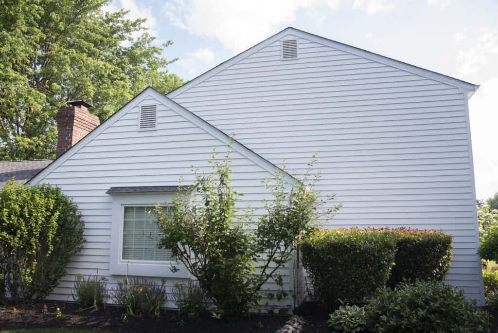 A white house with horizontal siding, two intersecting gable roofs, and a large window partly covered by green shrubs and bushes, with trees and a blue sky in the background.