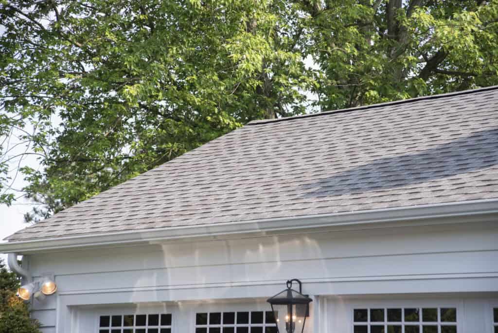 A gray shingled roof of a white house is shown, partially shaded by a large leafy tree overhead. A black outdoor lantern is mounted above windows on the exterior wall.