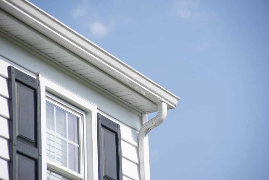 Close-up of the upper corner of a white house with black shutters, a window, and a white rain gutter against a clear blue sky.
