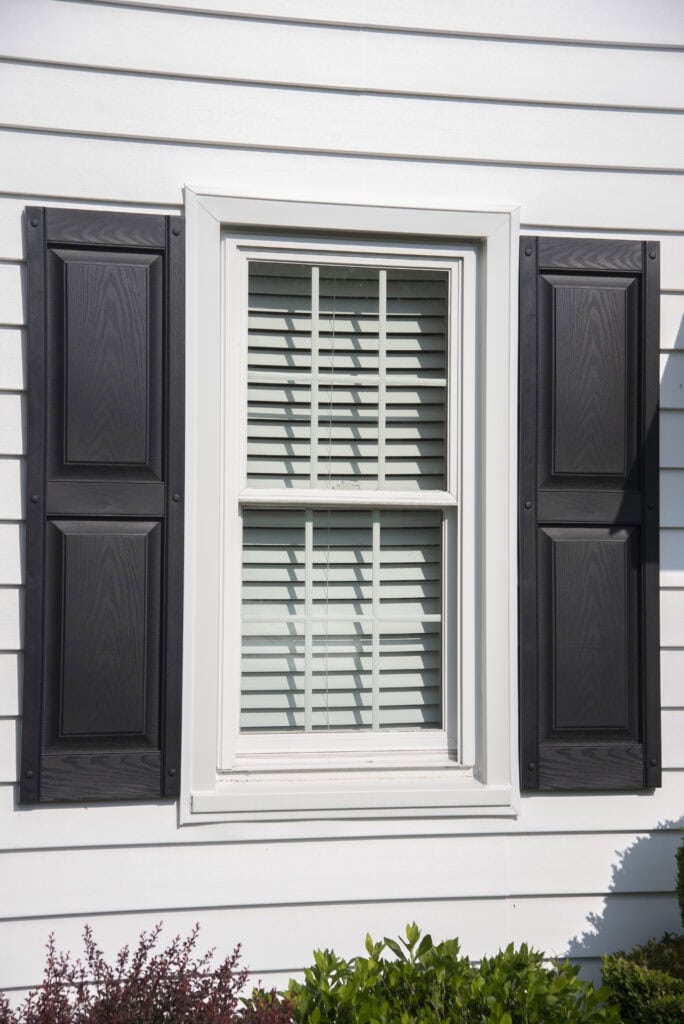 A white-framed window with closed blinds is set in white siding. Black shutters are on both sides of the window, and green shrubs are visible at the bottom.