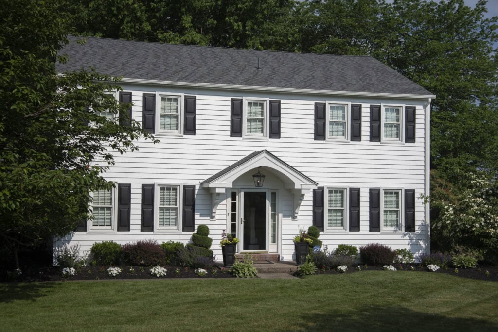 A two-story white house with black shutters, a dark gray roof, and a covered front entrance. The house is surrounded by green grass, shrubs, flowers, and trees in the background.