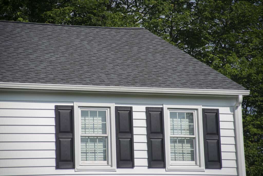 The image shows the upper section of a white house with two windows, each framed by black shutters, and a dark gray shingled roof. Green trees are visible in the background.