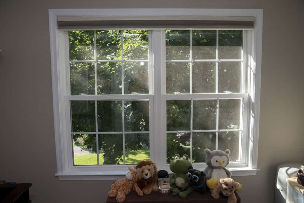 A sunlit window with white trim overlooks a green yard. In front of the window, several plush toys, including a lion, giraffe, frog, owl, and elephant, are arranged on a bench or shelf.