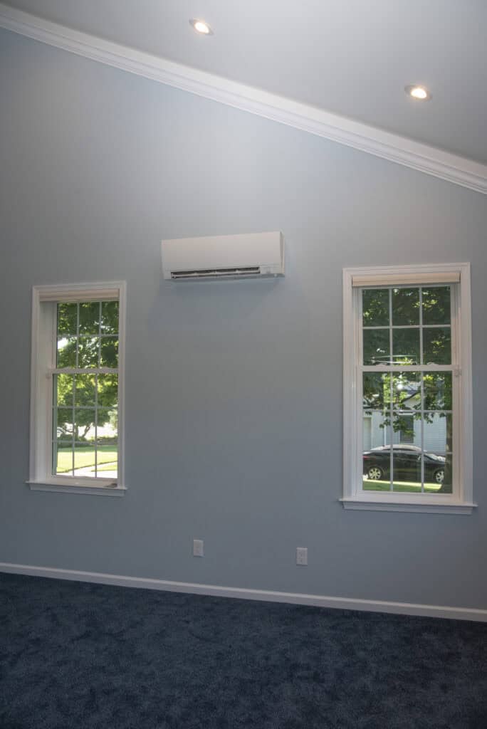 A light blue room with two windows, a wall-mounted air conditioning unit between them, blue carpet flooring, and recessed ceiling lights. Trees and a car are visible outside the windows.