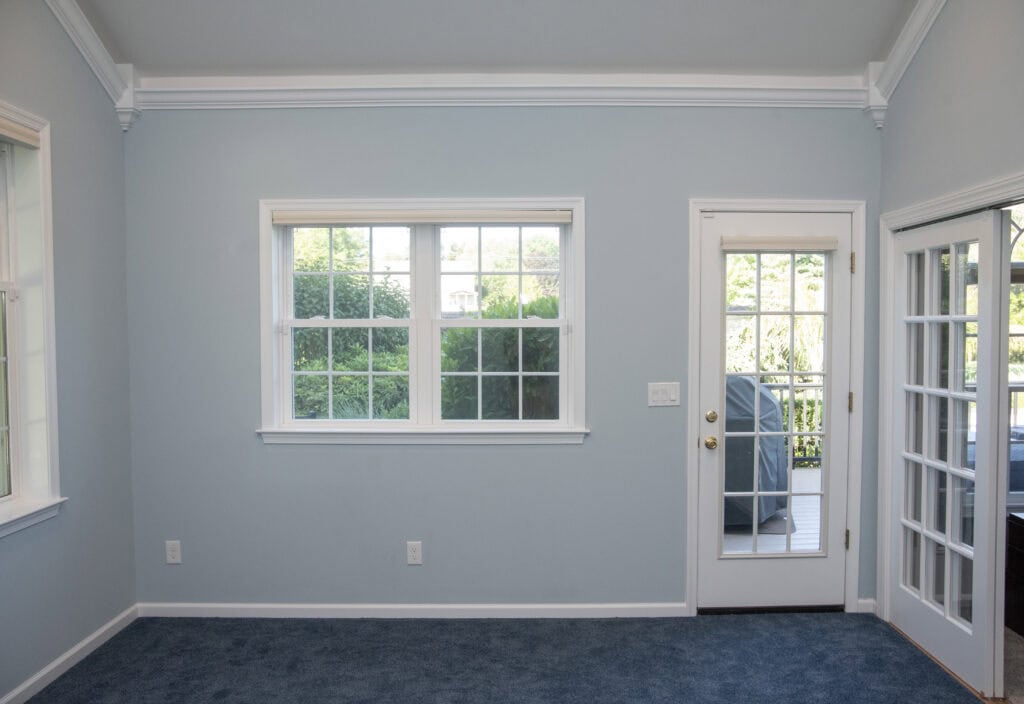 A bright room with light blue walls, a blue carpet, white trim, a large window, a glass door with a brass knob, and French doors leading to a porch with greenery outside.
