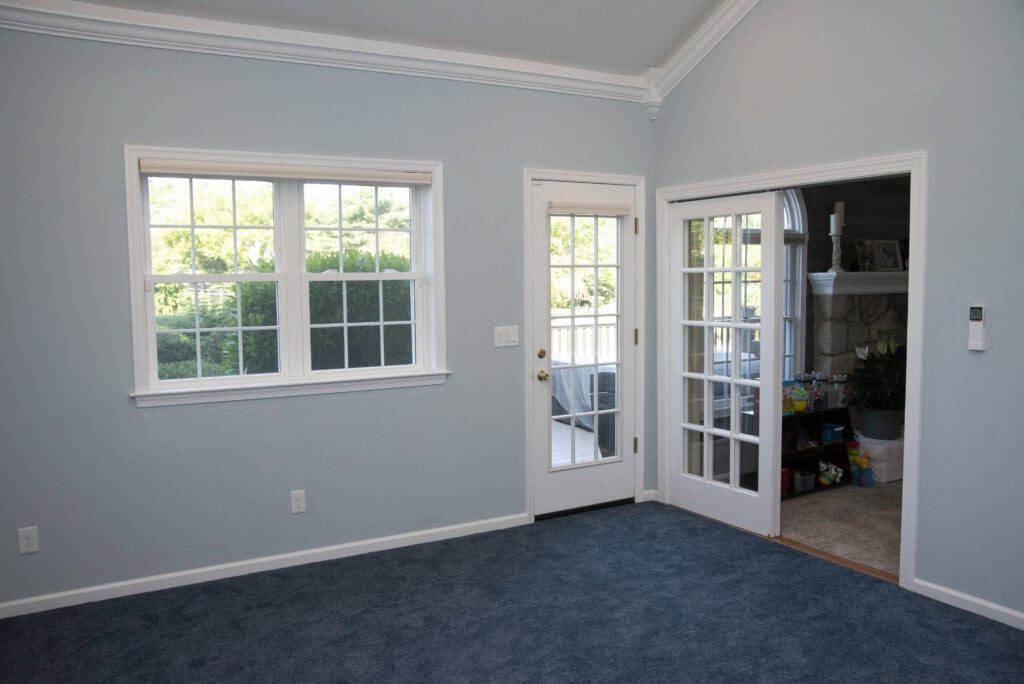 A bright room with light blue walls, dark blue carpet, two windows, a door with glass panes, and an open doorway leading to another room with a fireplace and toys visible.