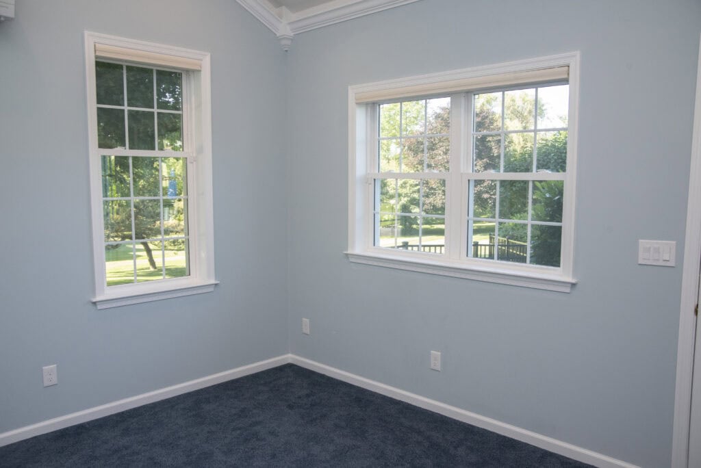 A corner of a room with light blue walls, white trim, and blue carpet, featuring two large windows letting in natural light and showing green trees and grass outside.