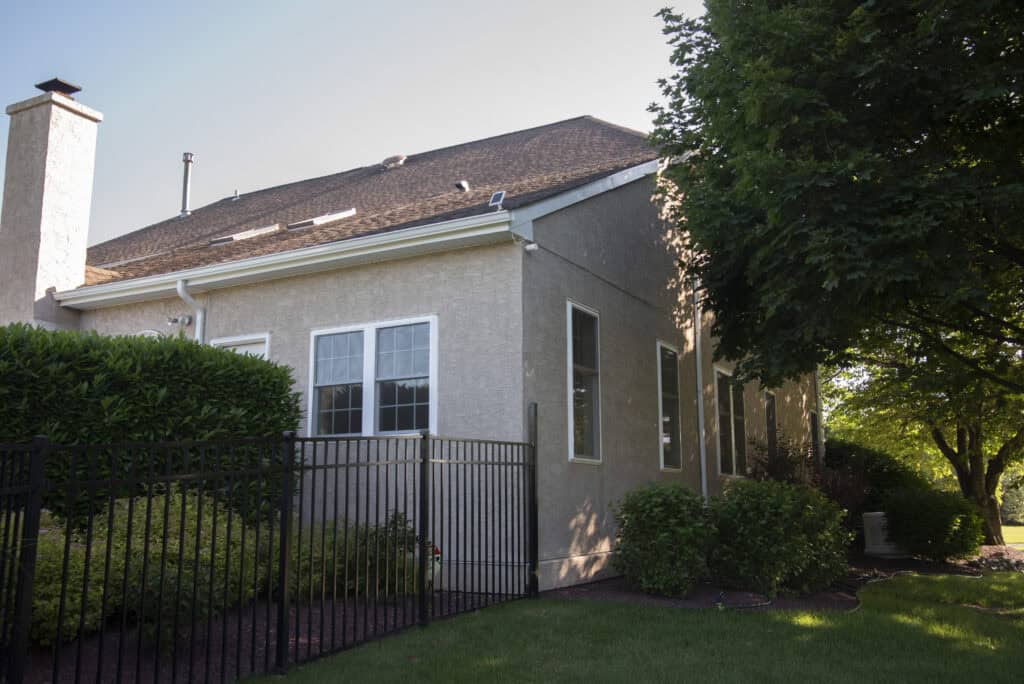 A beige house with several windows is bordered by a black metal fence and surrounded by green bushes, trees, and a well-maintained lawn on a sunny day.