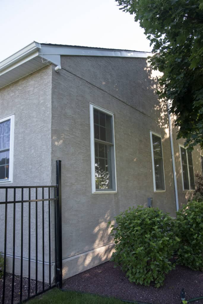 A beige stucco house with tall, narrow windows and white trim. A black metal fence and green shrubs are in the foreground, with a tree partially shading the building.