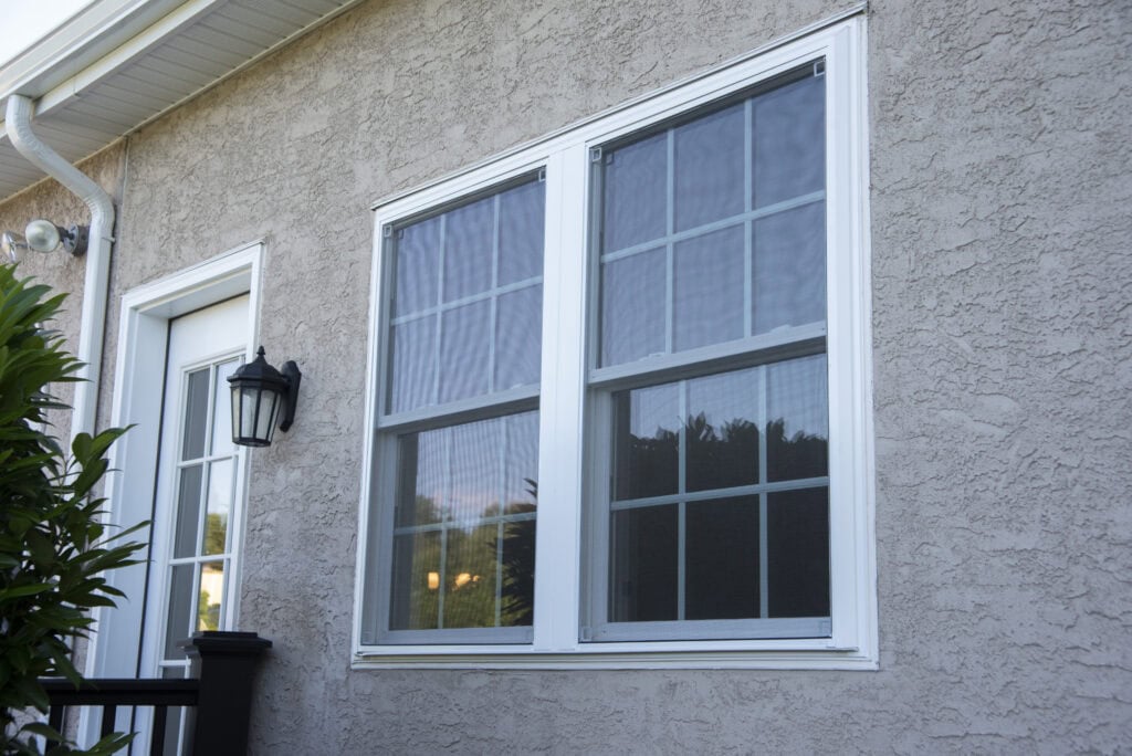 A close-up of a beige stucco house exterior featuring a white-framed window with grid panes and a nearby white door with glass panels. A black lantern-style light fixture is mounted beside the door. Green foliage is visible on the left.