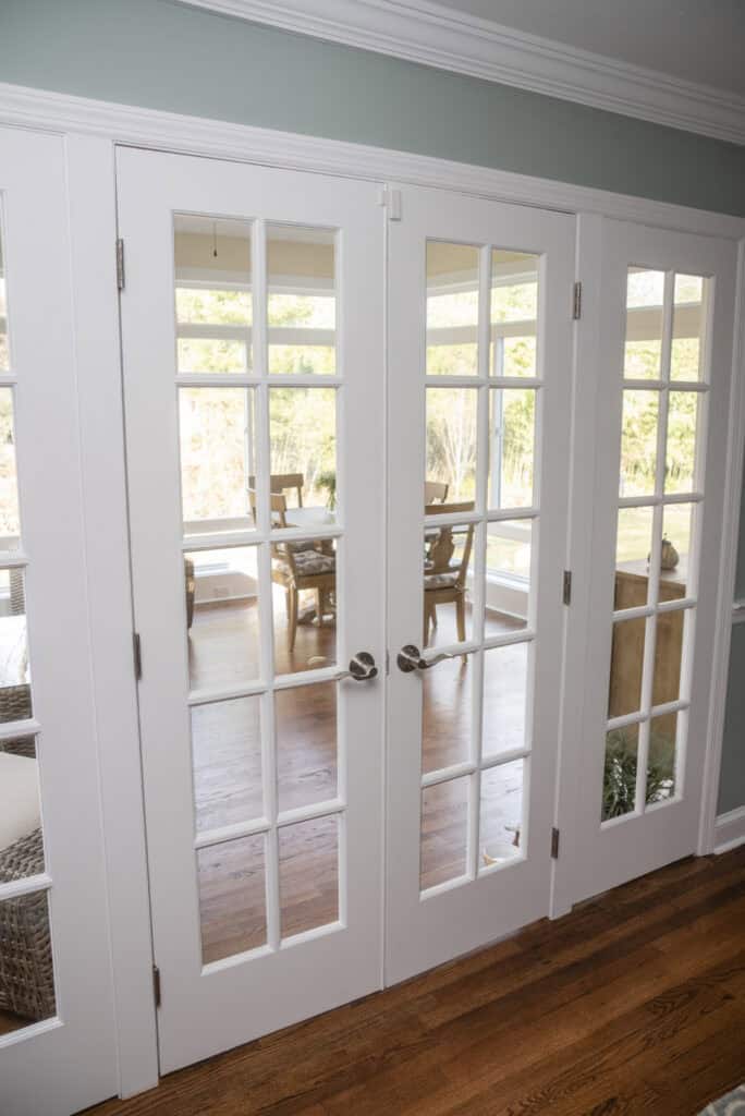 White French doors with glass panes lead to a sunlit room with wooden floors and a dining table. The room beyond is bright, with large windows showing an outdoor view with trees.