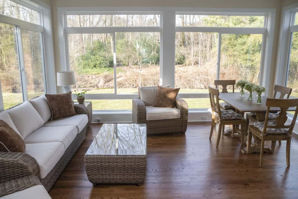 A bright sunroom with large windows, wicker furniture with white cushions, a wooden coffee table, and a dining table with six chairs, overlooking a green outdoor landscape.