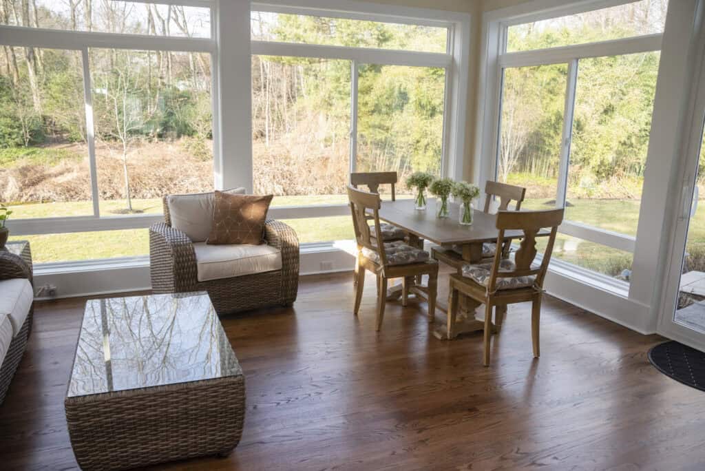 A bright sunroom with large windows, a wooden dining table set for four with small vases of flowers, a wicker armchair with a cushion, and a glass-topped wicker coffee table on hardwood floors.
