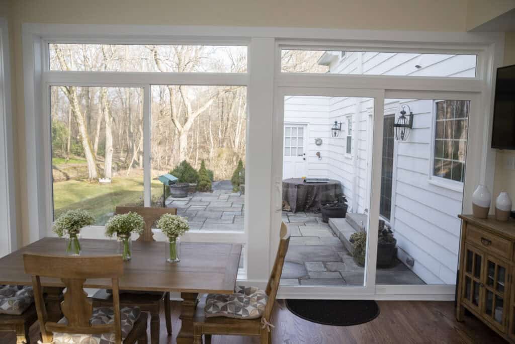 A dining table with four chairs and vases of white flowers sits by large windows, showing a view of a patio and a yard with leafless trees and grass outside. The interior has light walls and wooden floors.