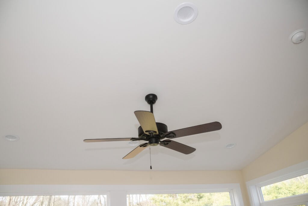 A ceiling fan with five dark blades is mounted to a white ceiling in a bright room with large windows and recessed lighting fixtures.