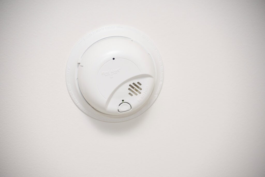 A white smoke detector mounted on a plain white ceiling. The device has a test button and vents on its face, and is centered in the image.