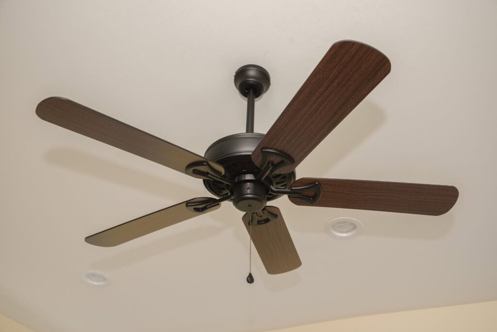 A dark brown ceiling fan with five wooden blades and a pull chain hangs from a white ceiling. Two recessed ceiling lights are visible in the background.