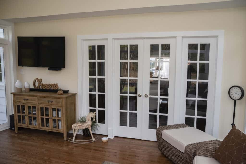 A cozy living room with wood floors, a beige couch, a small cabinet with decor, a mounted TV, a “Cheers” sign, a decorative rocking horse, a floor clock, and white-framed double French doors.