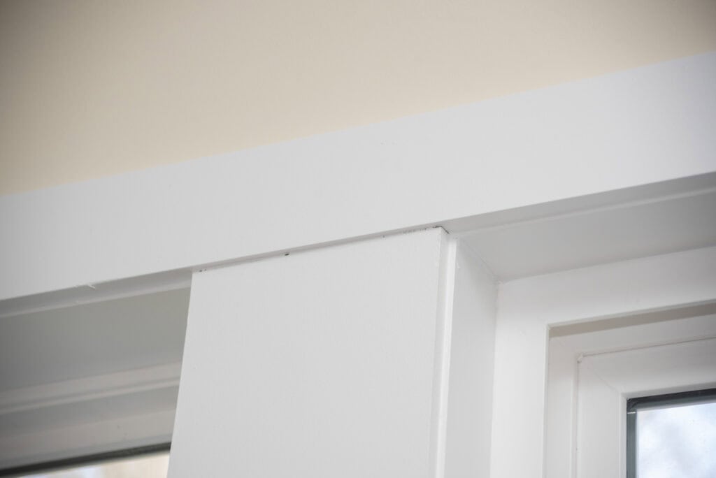 Close-up of the top corner of a white door frame and wall, showing clean lines and smooth surfaces with a window nearby. The image highlights the joint where the frame meets the wall.