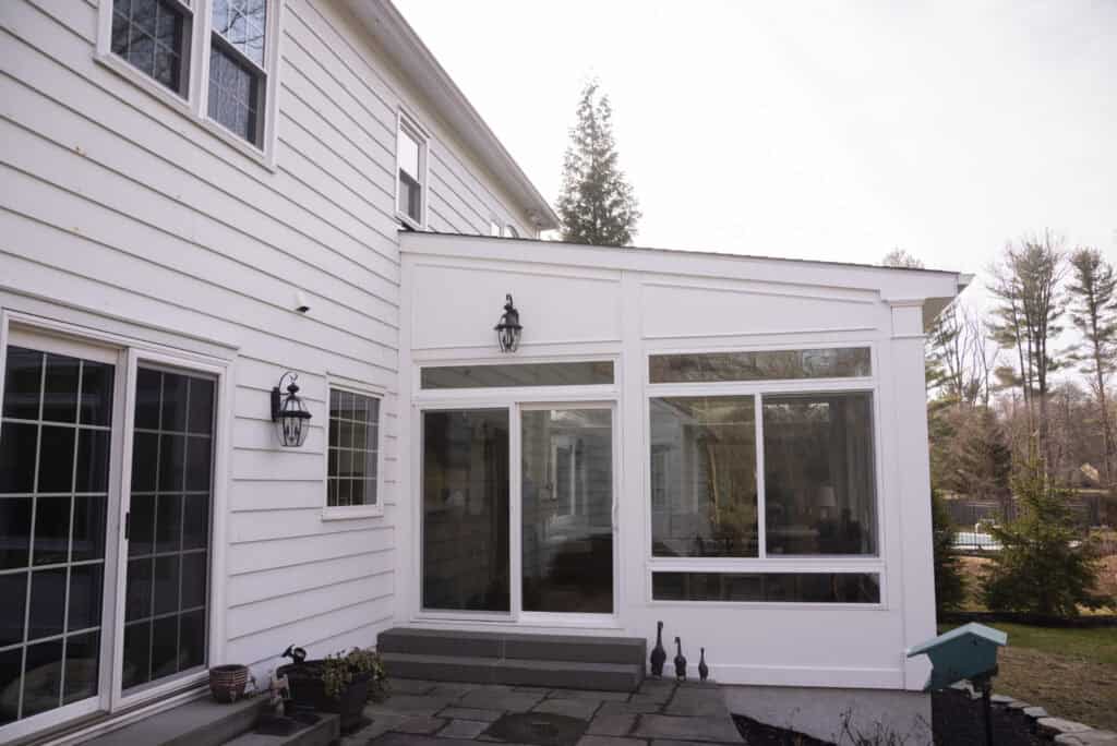 A white sunroom with large windows and sliding glass doors extends from the back of a house. Stone steps lead to a patio, and there are outdoor lights mounted on the house exterior. Trees are visible in the background.