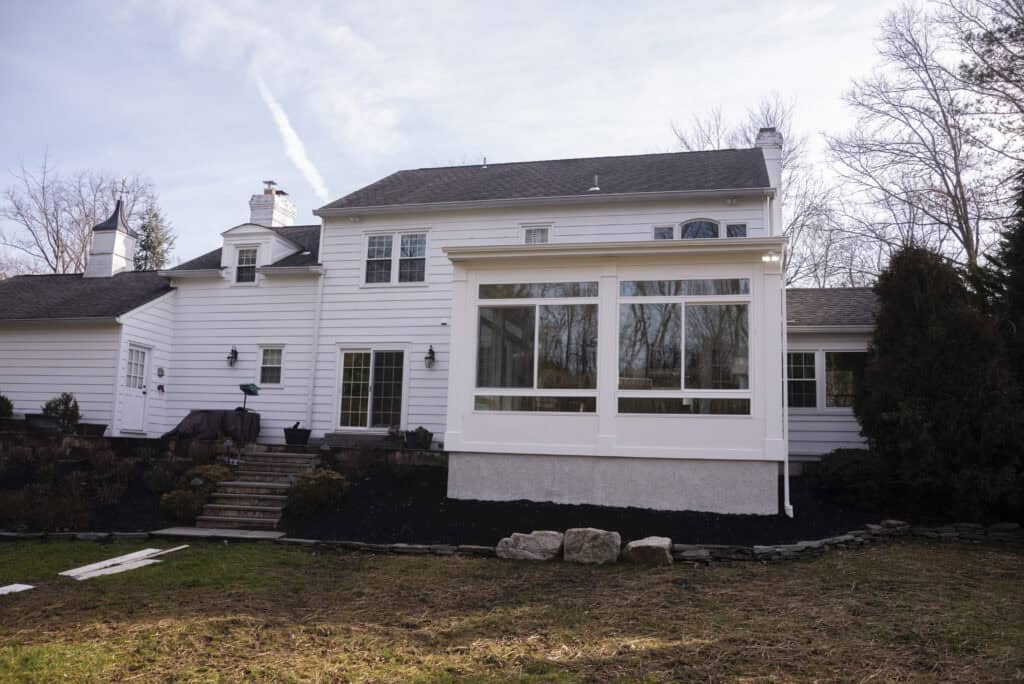 A large white two-story house with a modern sunroom extension featuring large windows. The yard has patches of bare soil, stone steps, and some landscaping with rocks and shrubs. Trees without leaves are in the background.