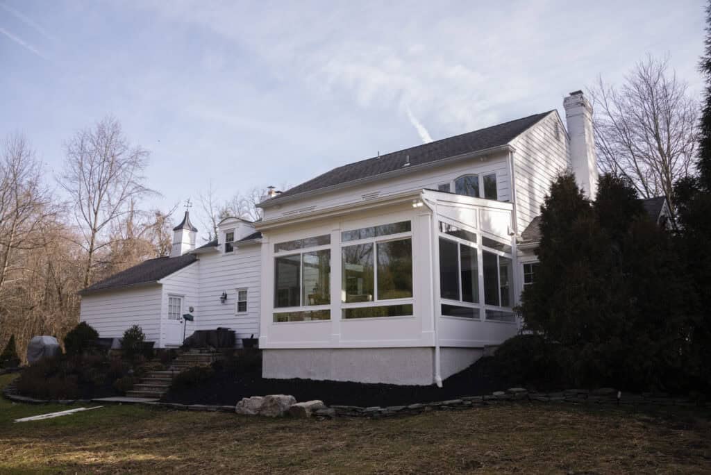 A white two-story house with large windows on a sunroom extension, surrounded by trees and shrubs, under a partly cloudy sky.