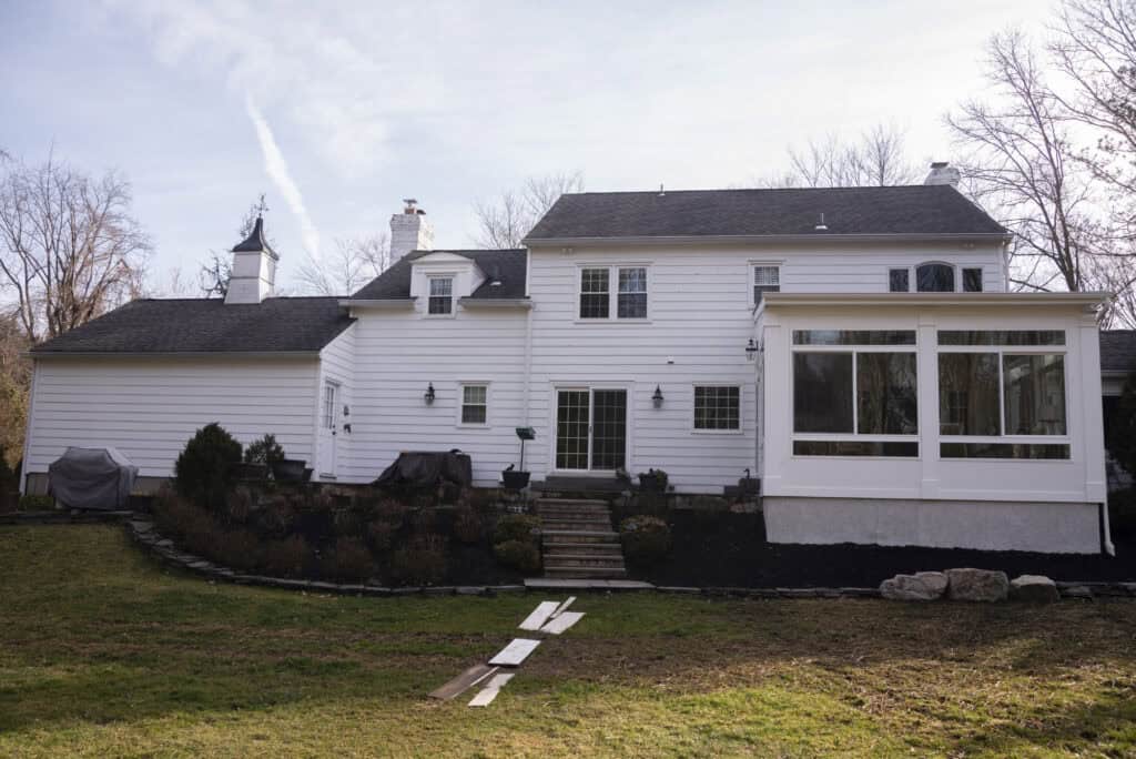 A large white two-story house with several windows and a sunroom extension, surrounded by bare trees and a grassy backyard with a stone path and landscaping.
