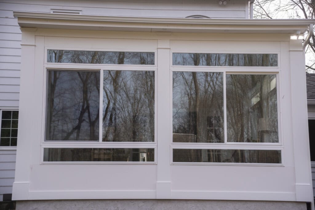 A white enclosed porch with large sliding glass windows reflects leafless trees and parts of a neighboring house on a cloudy day.