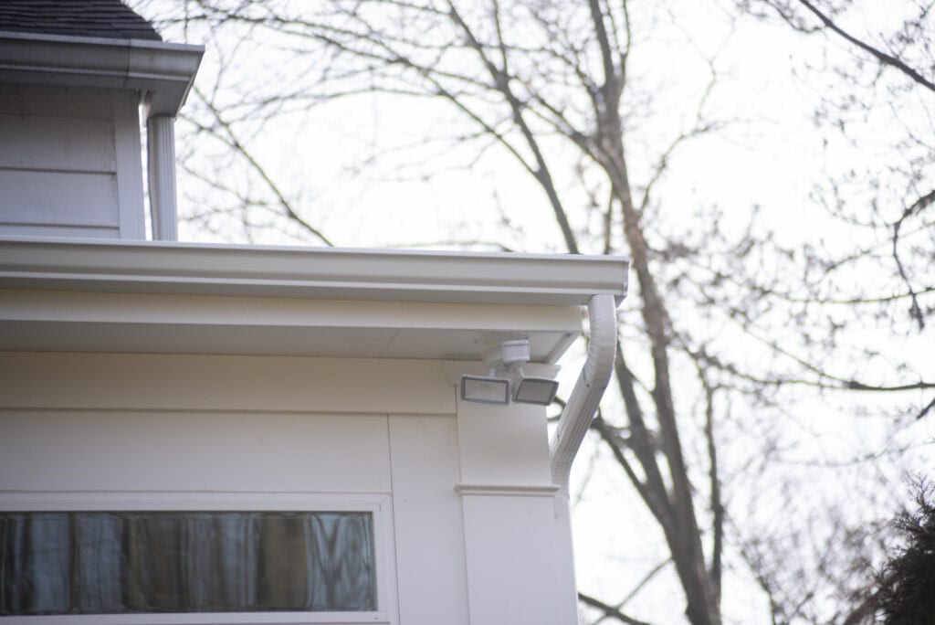 A close-up of the corner of a white house shows a security light mounted near the roof, with leafless trees and branches in the blurred background.