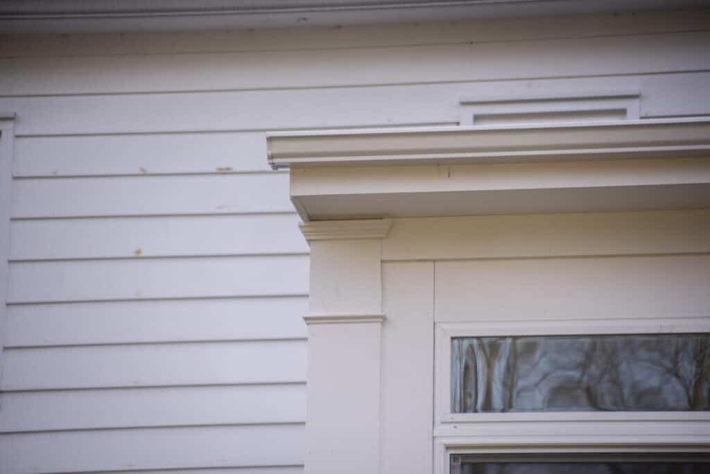 Close-up of a white house exterior featuring horizontal siding, the upper part of a doorframe with decorative trim, and the bottom edge of a window above, reflecting some tree branches.