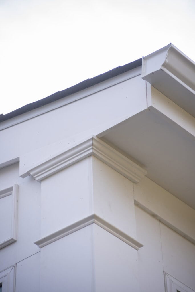 Close-up of the corner of a white modern building with decorative molding and a visible roof edge, photographed from a low angle against a bright sky.