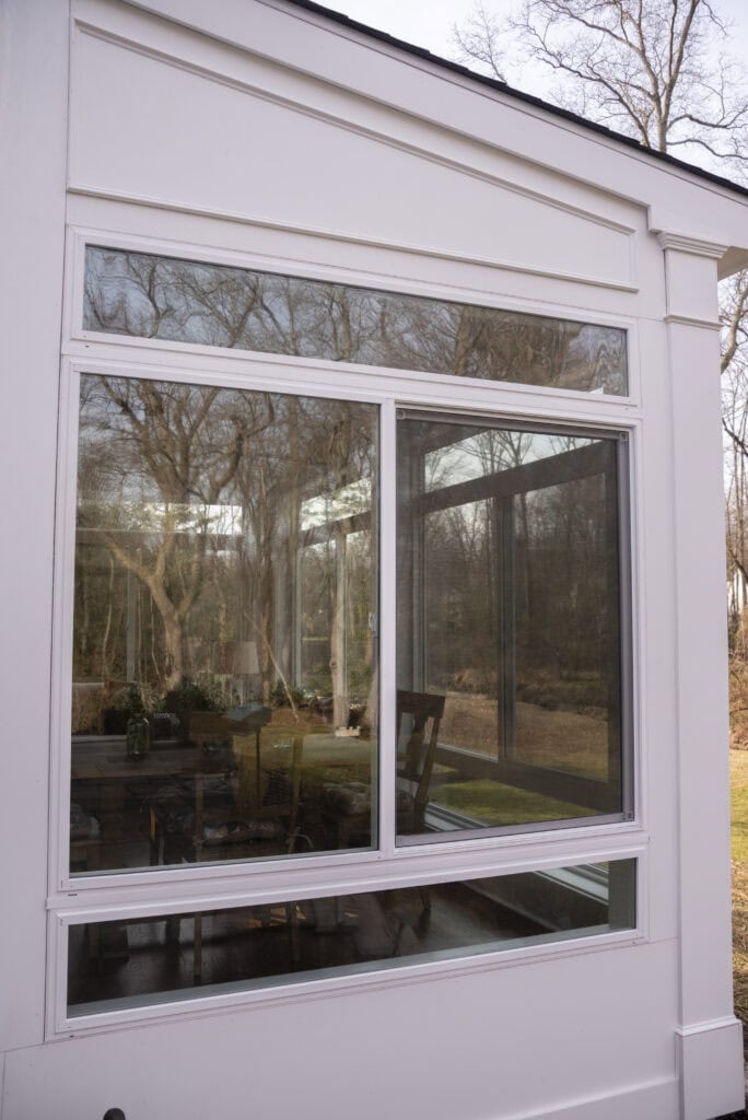 A large glass window reflects leafless trees and a cloudy sky; inside, a table, chairs, and plants are visible, suggesting a cozy indoor seating area with a view of the outdoors.