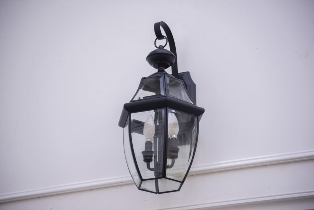 A black metal and glass outdoor lantern with two visible light bulbs is mounted on a white wall with decorative molding.