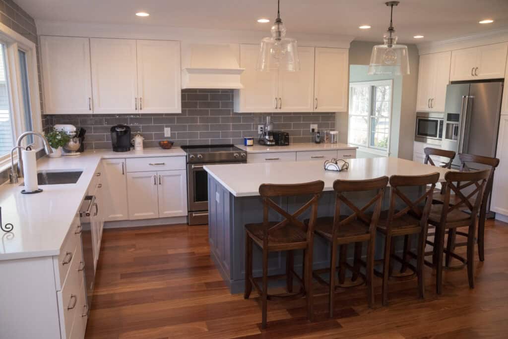 Modern kitchen with white cabinets, gray tile backsplash, stainless steel appliances, and a large island with four wooden chairs. Pendant lights hang above the island; wooden floors and ample natural light complete the space.