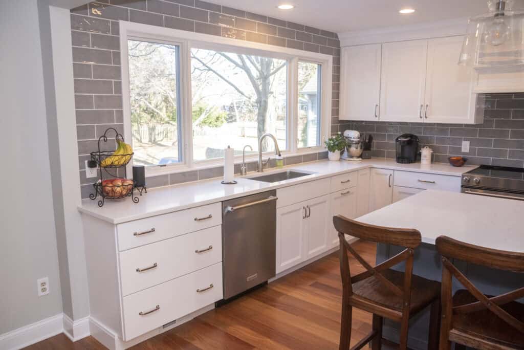 Bright, modern kitchen with white cabinets, gray subway tile backsplash, stainless steel appliances, large window over the sink, fruit basket on the counter, and two wooden barstools at a white island.