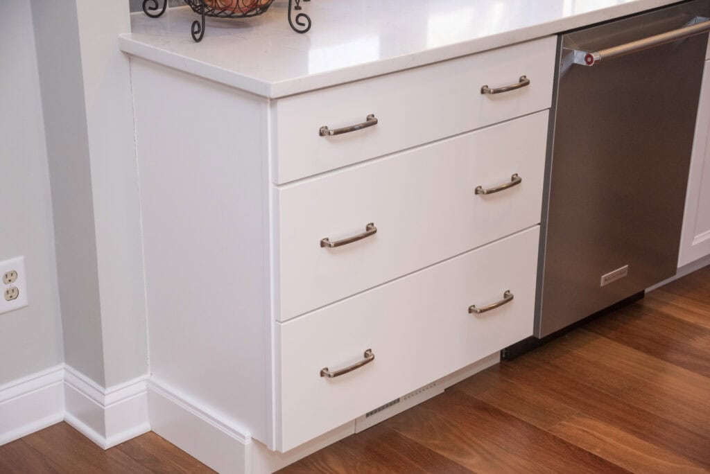 White kitchen cabinets with three drawers, silver handles, and a white countertop next to a stainless steel dishwasher. The cabinets sit on a wood floor, and a fruit bowl is partially visible on the counter.