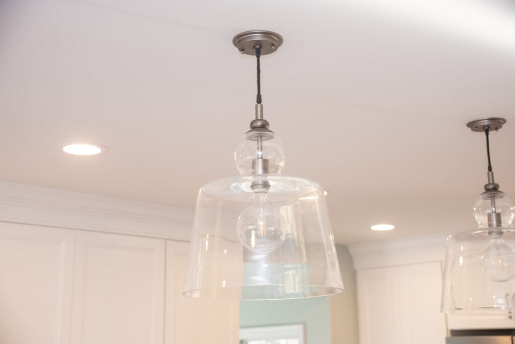 A clear glass pendant light fixture with a metal base hangs from a white ceiling in a modern kitchen with white cabinets and recessed lighting.