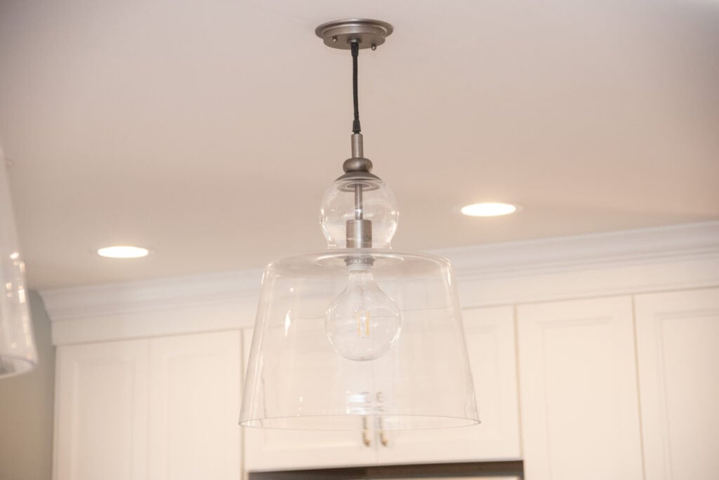 A modern pendant light fixture with a clear glass shade and exposed bulb hangs from the ceiling in a room with recessed lighting and white cabinets.