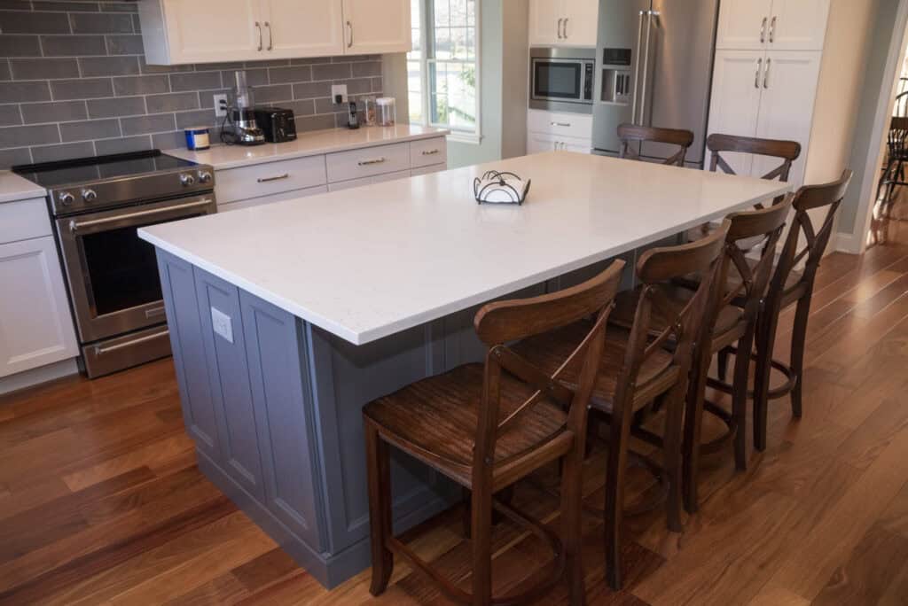 A modern kitchen with a large white island, four wooden chairs, stainless steel appliances, white cabinets, and hardwood floors. A window lets in natural light.
