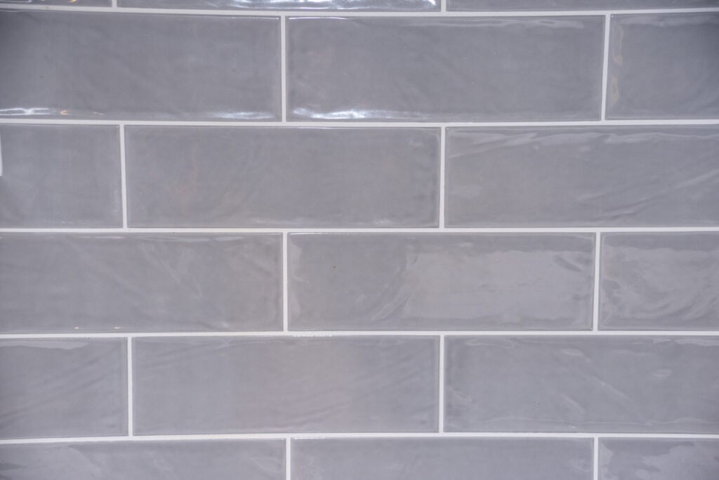 A close-up view of a wall with glossy, rectangular light gray tiles arranged in a horizontal brick pattern with white grout lines.