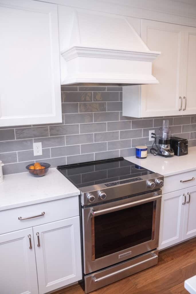 A modern kitchen features a stainless steel electric stove with a glass cooktop, white cabinets, gray subway tile backsplash, and small appliances on the countertop. A bowl of oranges and a canister sit nearby.