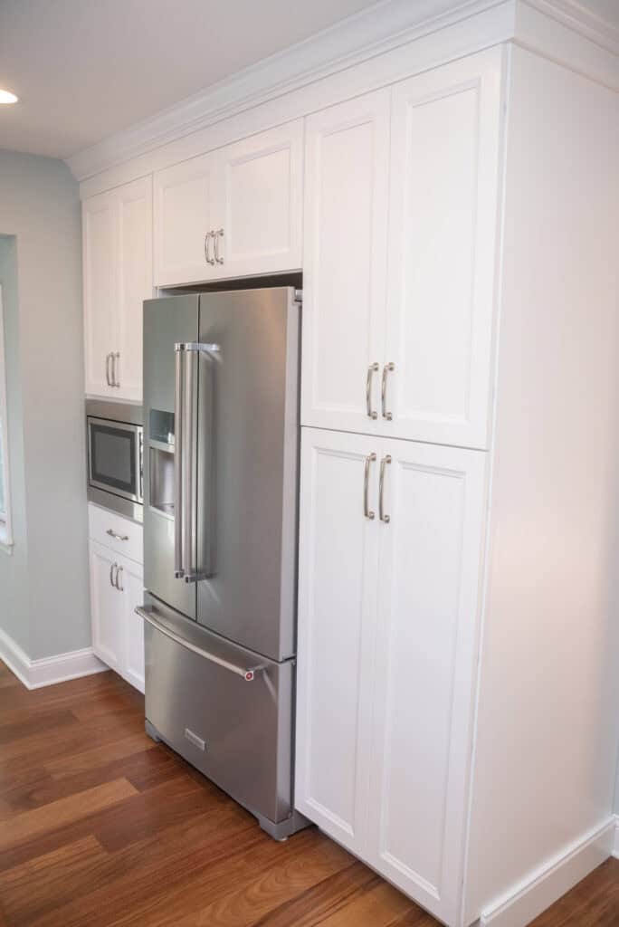 A modern kitchen features a stainless steel French door refrigerator, built-in microwave, and white cabinets with silver handles, all set against a light blue wall and wood flooring.