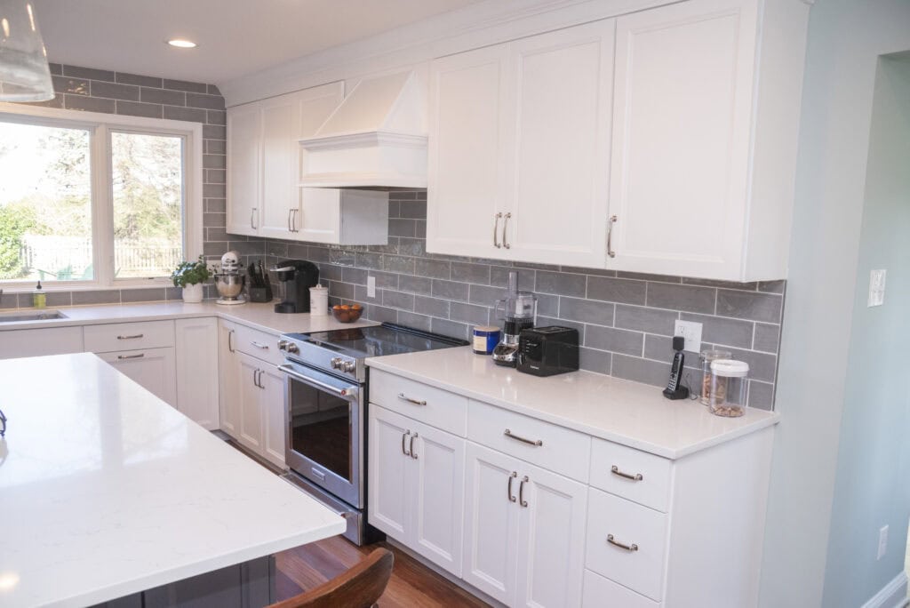 Modern kitchen with white cabinets, gray subway tile backsplash, white countertops, stainless steel appliances, and a large window letting in natural light. Kitchen gadgets and small decor items are on the countertop.