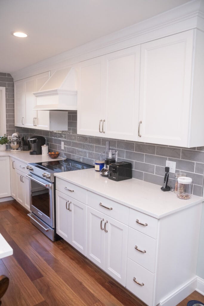 Modern kitchen with white cabinets, stainless steel appliances, gray subway tile backsplash, and wooden floor. Small appliances, a bowl, and a plant are on the white countertop.