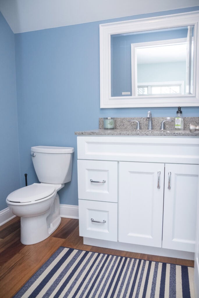 A small bathroom with blue walls, a white toilet, a white vanity with a granite countertop, a sink, soap dispenser, candle, large mirror, and a blue and white striped rug on hardwood flooring.