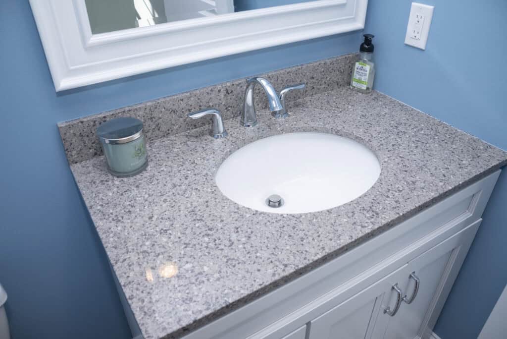 A bathroom sink with a speckled gray countertop, silver faucet, a green candle, and hand soap dispenser. The vanity is white with silver handles, against a light blue wall with a mirror above.