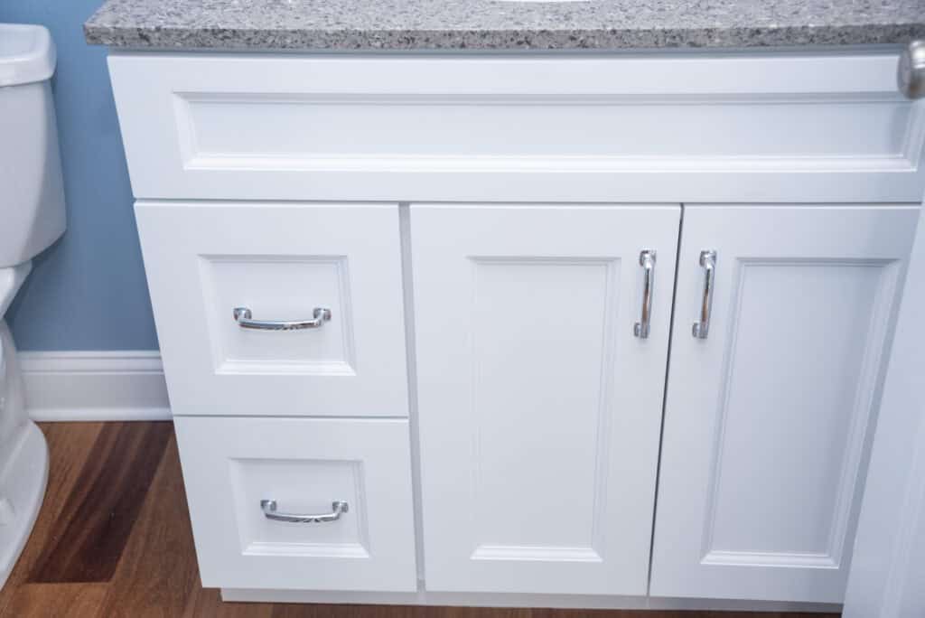 White bathroom vanity with silver handles, featuring two drawers on the left and two cabinet doors on the right, topped with a gray speckled countertop; part of a toilet and wooden floor are visible.