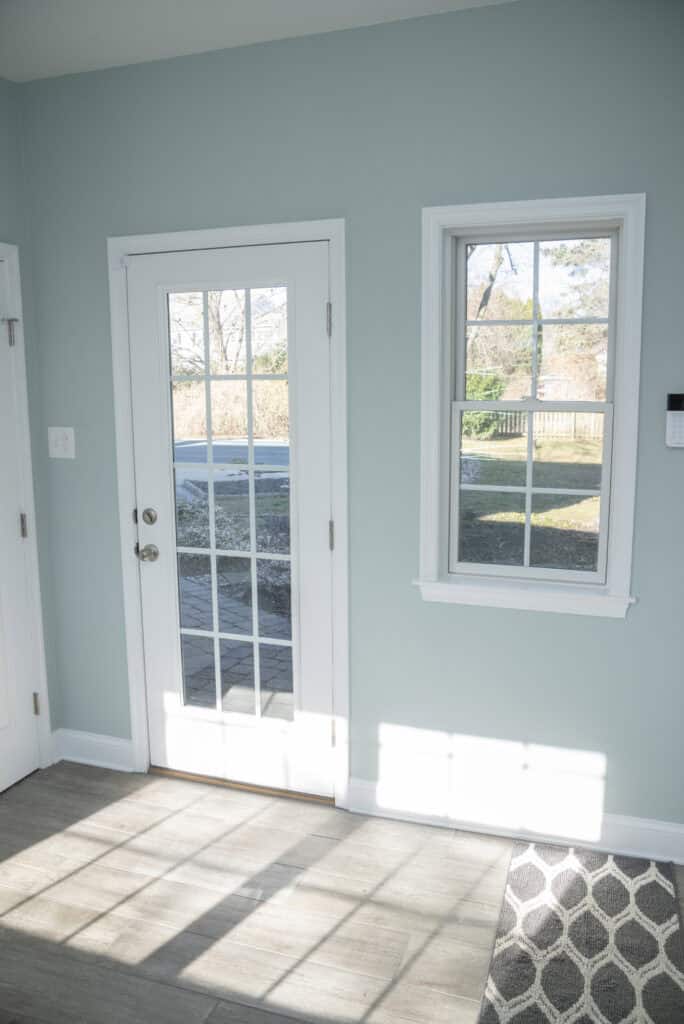 A sunlit room with a glass-paneled white door and a window next to it, both showing an outdoor view. Sunlight casts shadows on the light wood floor and part of a gray patterned rug.