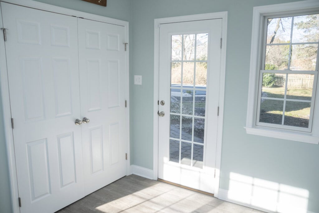 A bright entryway with a glass-paneled door, two white double-door closets, and a window, all letting in natural sunlight onto light-colored tile flooring. Outdoor trees and yard are visible through the glass.
